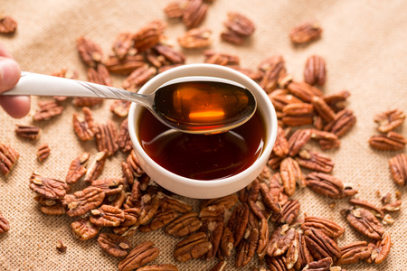 Maple Syrup in Bowl with Pecans Close Up. a close up view of a bowl of maple syrup with a spoon dipped in and pulling some out surrounded by pecans on a textured brown clothの写真素材