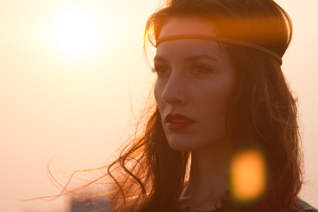Portrait of a hippie woman with headband looking far away at sunset with windy hair. Shot with anamorphic flare. Vintage looking hippie girl looking at distance. Closeupの写真素材