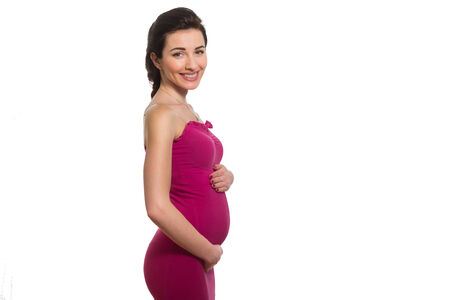 Young caucasian smiling pregnant lady in pink dress touching belly in profile isolated on white backgroundの写真素材