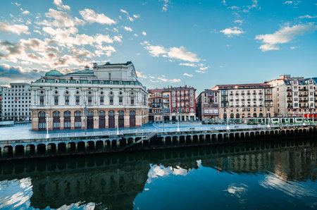 Arriaga Theater building standing on the waterfront reflecting in the calm Nervion River with traditional architecture and a modern tram passing by on a bright day. Bilbao Spain October 21th 2025のeditorial素材