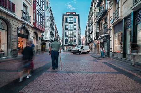 Elderly man with a cane walking on a brick pavement street, with blurred people creating a sense of urban movement and daily life in Bilbao, Spain. Bilbao Spain October 20th 2025のeditorial素材