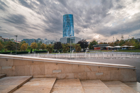 Iberdrola Tower rising prominently in Bilbao's urban landscape, showcasing contemporary architecture amidst a park with fountains and warm toned stairs leading into the sceneのeditorial素材