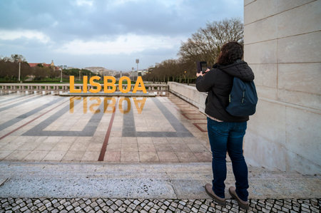 Tourist standing on cobbled ground, capturing the yellow Lisboa sign reflecting in the water feature...
