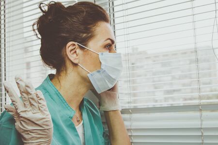 Stress woman Doctor in medical mask, gloves and doctors suit talking on a cell phone near the windowの写真素材