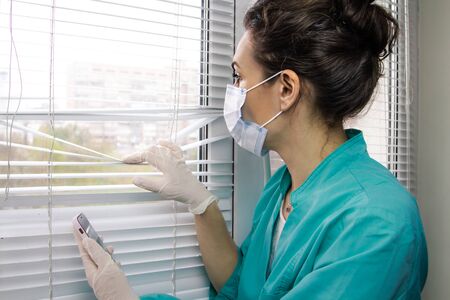 Tired exhausted doctor from Italy hospital in a medical surgical mask and doctor's uniform looks out the window through the ajar blinds.の写真素材