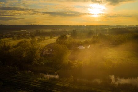 Golden orange mist over the village. Dramatic morning landscape with fog, river and meadow in the golden hour at dawn.の写真素材