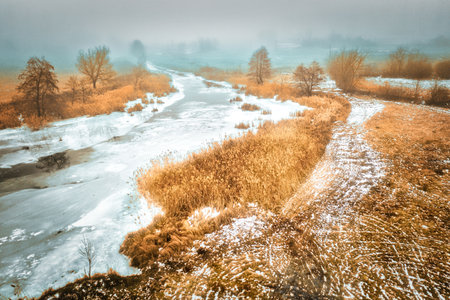 Off-season on a melting river - thawed patches on ice among yellow dry grass and trees.の写真素材