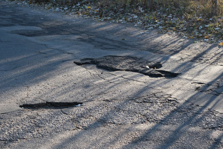A detailed closeup of a damaged asphalt road showing visible cracks and patched areas, beautifully illuminated by soft, natural light, effectively highlighting various road maintenance issuesの写真素材