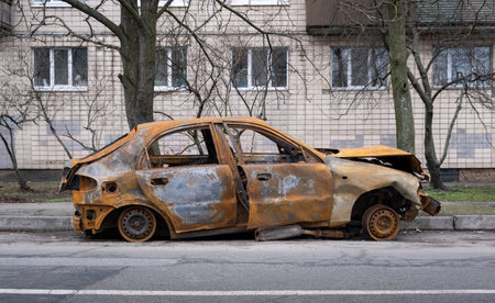 A detailed closeup view of an abandoned car, which is rustcovered and leaning against a cracked sidewalk, set beneath a backdrop of bare trees, powerfully showcasing urban decay and neglectの写真素材