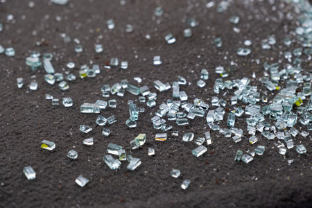 A detailed closeup view of tiny shards of broken glass scattered across a dark, textured surface, effectively capturing the beautiful reflective qualities and shimmering aspects of the glass piecesの写真素材