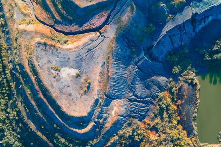 Stunning aerial drone view of massive open-pit mine or quarry showing terraced earth in deep blues and purples, contrasting with vibrant autumn vegetation and artificial lakes. Industrial landscape.の写真素材
