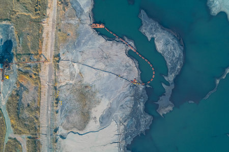 Aerial abstract composition of a mining tailings pond. A dredge pipe snakes across grey sludge and...