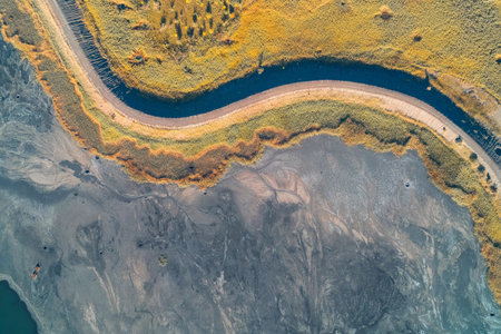 Abstract aerial geometry of an S-curve road path separating environmental zones. Yellow reeds border a dark blue channel, sharply divided from the gray industrial tailings below land stress.の写真素材