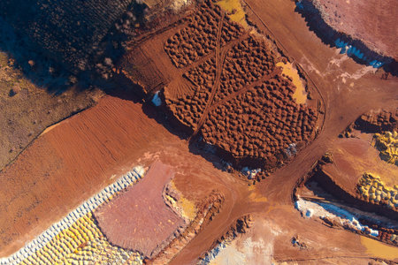 Abstract drone shot of an open-pit mine featuring organized mounds of red clay and bright yellow mineral deposits. The textured earth piles and muddy pools highlight industrial extraction.の写真素材
