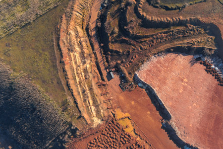 Abstract drone shot of an open-pit mine. The red clay terraces, white rock piles, and a small truck highlight the massive scale of industrial extraction and raw material development.の写真素材
