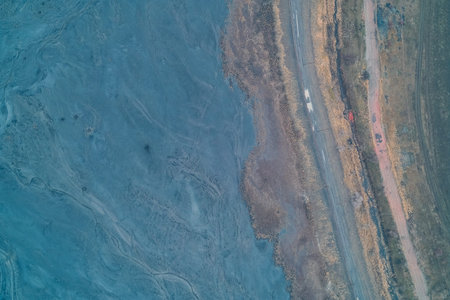 Aerial drone shot of a transport road separating a blue chemical sediment pond from dry fields. Top...