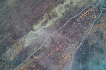 Winding dirt roads and vehicle tracks cutting through a massive scorched earth field. The terrain is scarred with red and black textures and faint earthworks in a war zone area. High-angle drone perspective.の写真素材