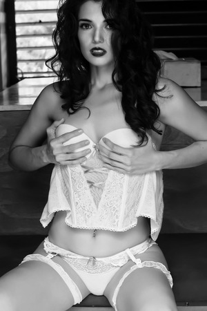 An image of a very attractive woman in white lingerie sitting on a bench in a room, adjusting her bodice, in a room with ambient light and fill flash, in an attitude of self-confidence and intimacy, color, vertical,の写真素材