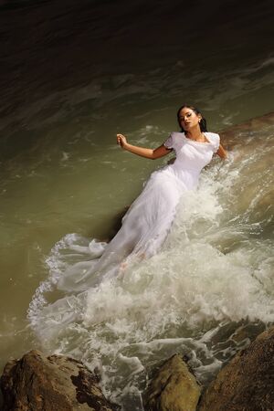 Photograph of a beautiful model dressed as a bride, perched on a rock, with the veil over her body, while the waves of the sea fall on her, in an attitude of confidence and serenity, with a background of the sea, dusk, color, verticalの写真素材