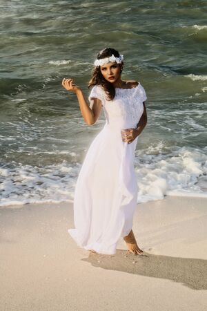 A photo of a beautiful young Latina woman model, dressed in a beautiful designer wedding dress and with headdress, with earrings, walking on the beach and with a very confident attitude and sensual relaxation, looking at the camera, with a sea bottom, colorの写真素材