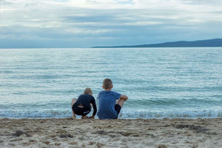 Two young brothers sitting on a beach in St Heliers, Auckland, New Zealand. The older brother is looking at the sea and an island on the other side and the younger brother is playing in the sand.の写真素材