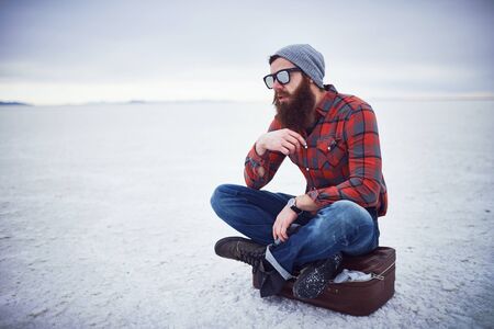 pensive contemplative hipster stroking awesome manly beard sitting on retro suitcase in salt flats all aloneの写真素材