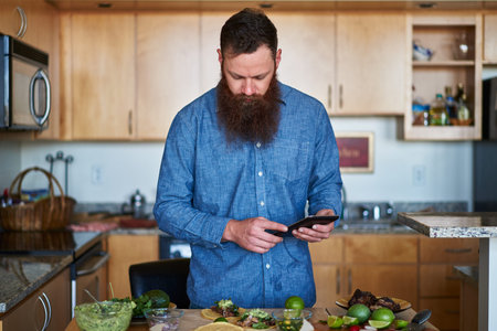 trendy man with cool beard using tablet or smartphone to look up taco recipe in kitchenの写真素材