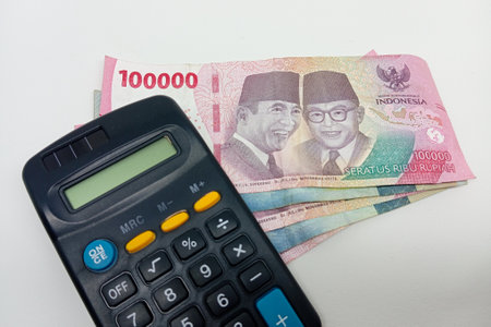 A dark-colored calculator with yellow and blue buttons is positioned next to a fan of Indonesian 100,000 Rupiah banknotes, featuring the portraits of Sukarno and Mohammad Hattaの写真素材