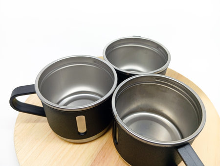 Three black stainless steel mugs arranged on a round wooden board, isolated on white background. Modern camping or kitchenware set.の写真素材