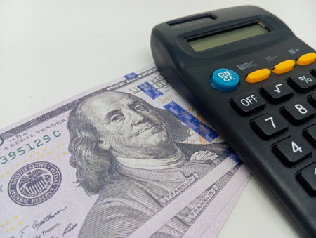 US $100 bills fanned out next to a dark-colored basic calculator, isolated on a clean white background, signifying financial calculation or budgetingの写真素材