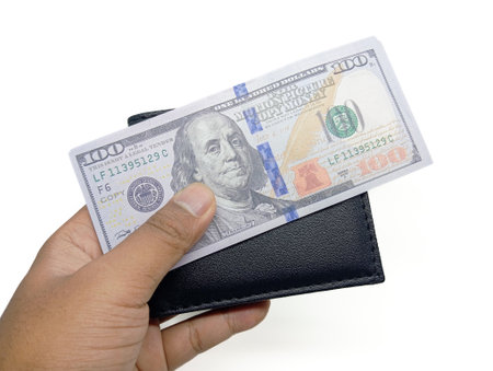 A human hand holds a black leather wallet from which a US 100 dollar bill is partially pulled out, against a clean white background.の写真素材