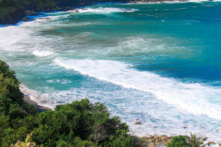 A high-angle view showcases a stunning tropical bay with turquoise waters, foamy white waves breaking on a sandy beach, and lush green coastline vegetation.の写真素材