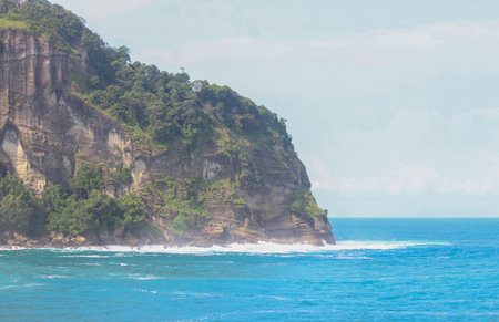 A majestic coastal cliff, covered in lush green vegetation, dramatically meets the vibrant turquoise ocean with white waves crashing at its base, under a bright sky.の写真素材