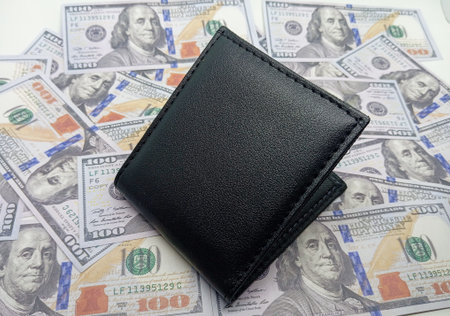 A sleek black leather wallet lies open on a scattered bed of US 100 dollar bills, symbolizing wealth...
