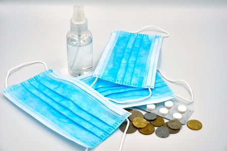 Three blue surgical face mask with bottle of sanitizer, coins and medicine pills on whiteの写真素材