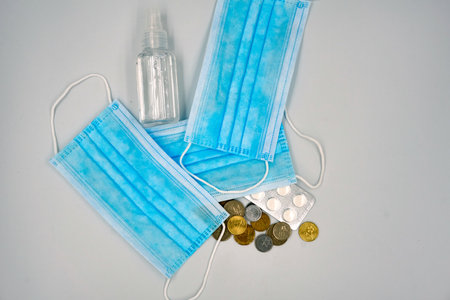 Three blue surgical face mask with bottle of sanitizer, coins and medicine pills on whiteの写真素材