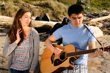 A young adult female watches a young adult male play a song on his guitar