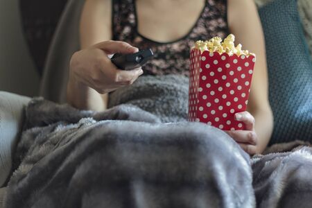 Woman wrapped in warm blanket holding tv remote control and holding box of popcornsの写真素材