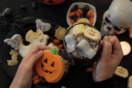 Top view of woman eating a pumpkin cookie and drinking spooky hot drink on table with halloween food and decorationの写真素材