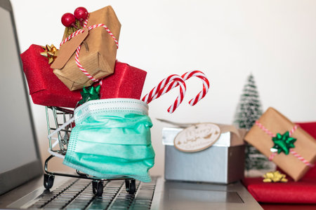 Shopping cart full of Christmas gifts and presents and protective face mask on laptop computer. Concept of online shopping at Christmas during covid-19 pandemicの写真素材