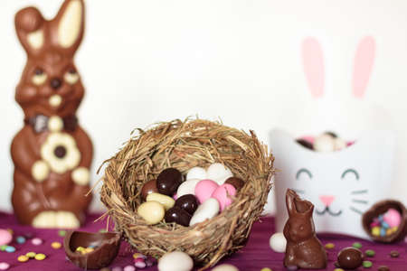 Bird nest with easter almonds, chocolate eggs, chocolate bunny and sweets on table against white background. Easter celebration conceptの写真素材