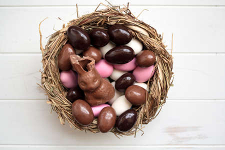 Top view of bird nest with chocolate bunny and sweet easter eggs on white wooden tableの写真素材