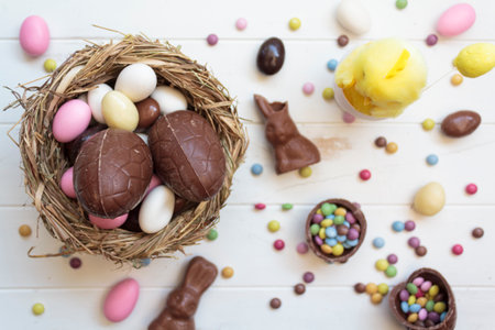 Top view of bird nest with chocolate easter eggs, chocolate bunny and sweets on white wooden tableの写真素材