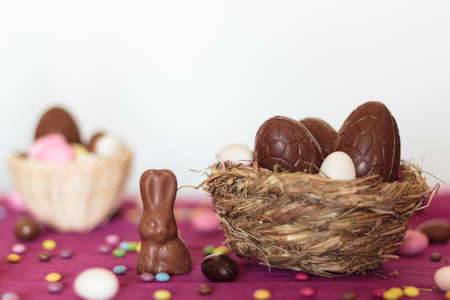 Bird nest with chocolate eggs and chocolate bunny and other easter sweets on table against white backgroundの写真素材