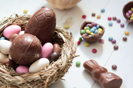 Chocolate eggs and easter almonds on bird nest, chocolate bunny and sweets on white wooden tableの写真素材