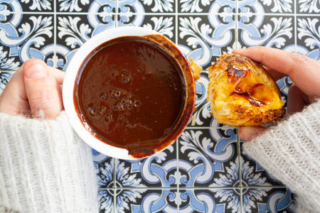 Top view of woman hands holding cup with hot chocolate and portuguese custard tart called pastel de nata on portuguese tiles backgroundの写真素材