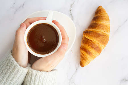 Top view of woman hands holding cup with hot chocolate drink and french croissant on marble tableの写真素材