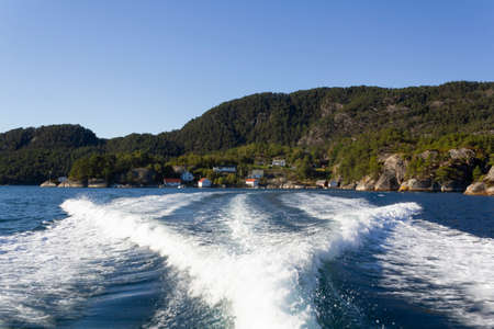 Trail on water surface of fast moving boat with norwegian houses on green forest mountains on the backgroundの写真素材