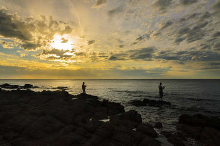 Silhouette two adult men fishing from rocks at sunsetの写真素材