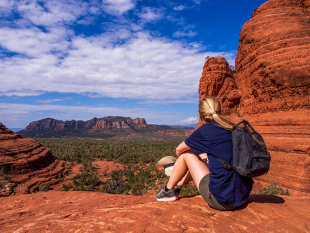 A woman takes a moment to admire the breathtaking views of Sedona, Arizona, while sitting on the red rocks wearing a blue shirt and carrying a backpack. The scenic red rock formations and stunning natural beauty of the area make it a popular destination for outdoor enthusiasts and nature lovers.の写真素材